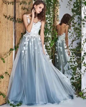 Bride in an ethereal ivory tulle gown with 3D floral appliqués, posing against a rustic backdrop with vines. The dress features an illusion neckline and delicate detailing, enhancing its fairytale-like appearance.