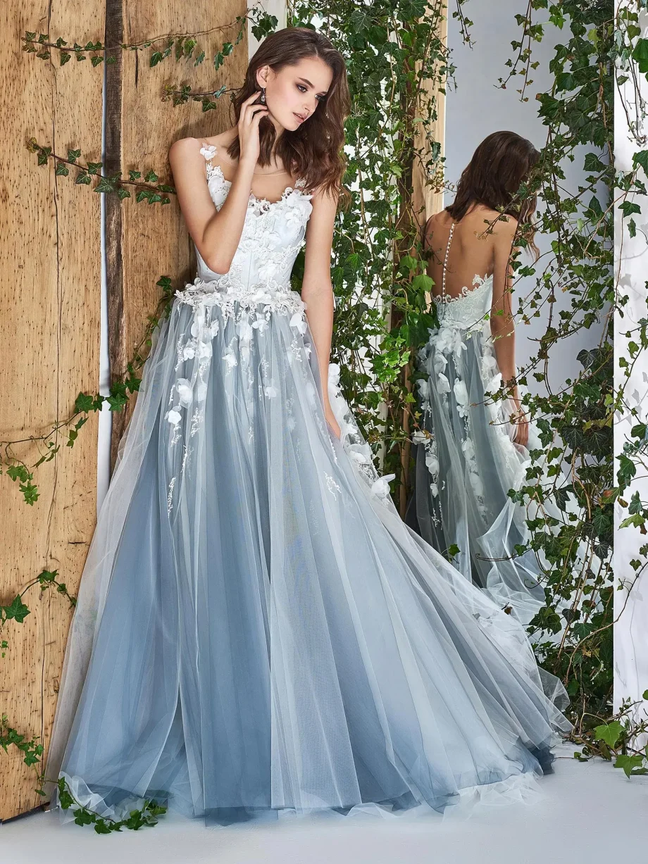 Bride in an ethereal ivory tulle gown with 3D floral appliqués, posing against a rustic backdrop with vines. The dress features an illusion neckline and delicate detailing, enhancing its fairytale-like appearance.