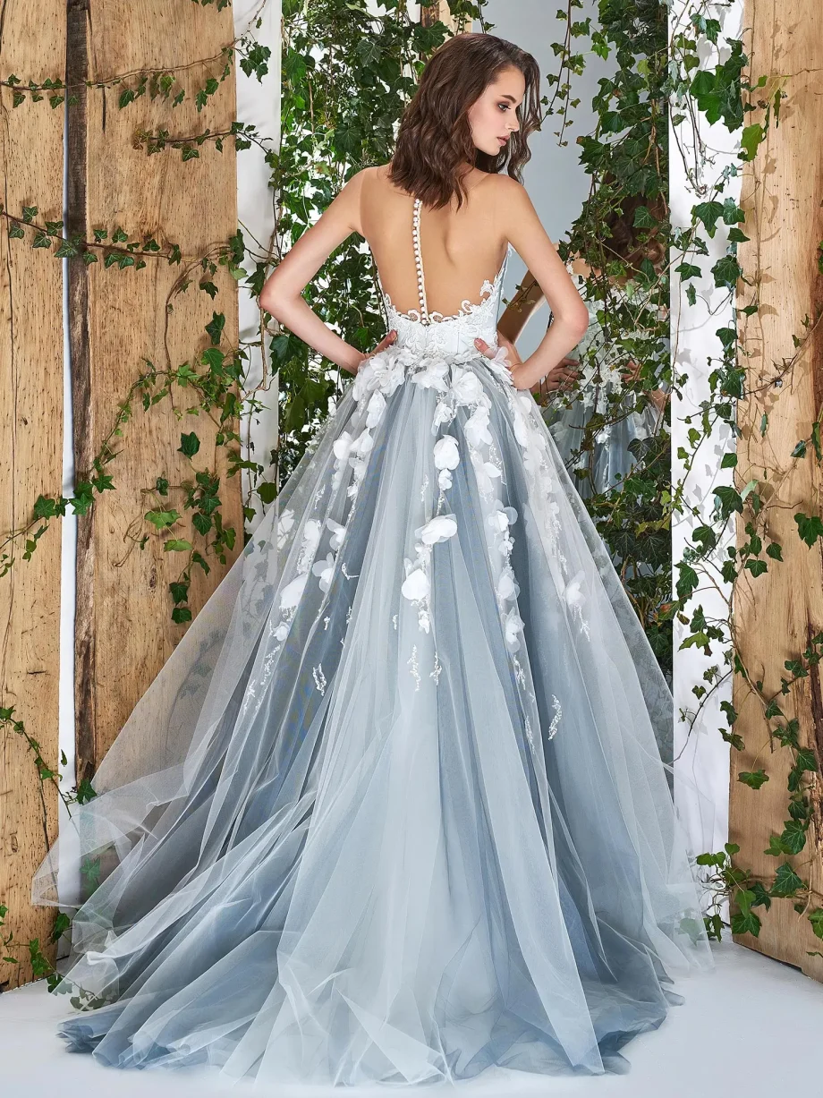 Back view of a bride in an ivory tulle wedding gown showcasing an illusion back with button details and layers of flowing tulle, set against a wooden and vine-covered backdrop for a natural, enchanting look.