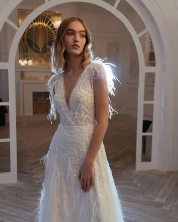 A bride in a dazzling V-neck wedding gown with feathers, part of the Papilio Boutique sample sale in Toronto. The gown features a deep V-neckline and is photographed in a luxurious room with white and gold decor.