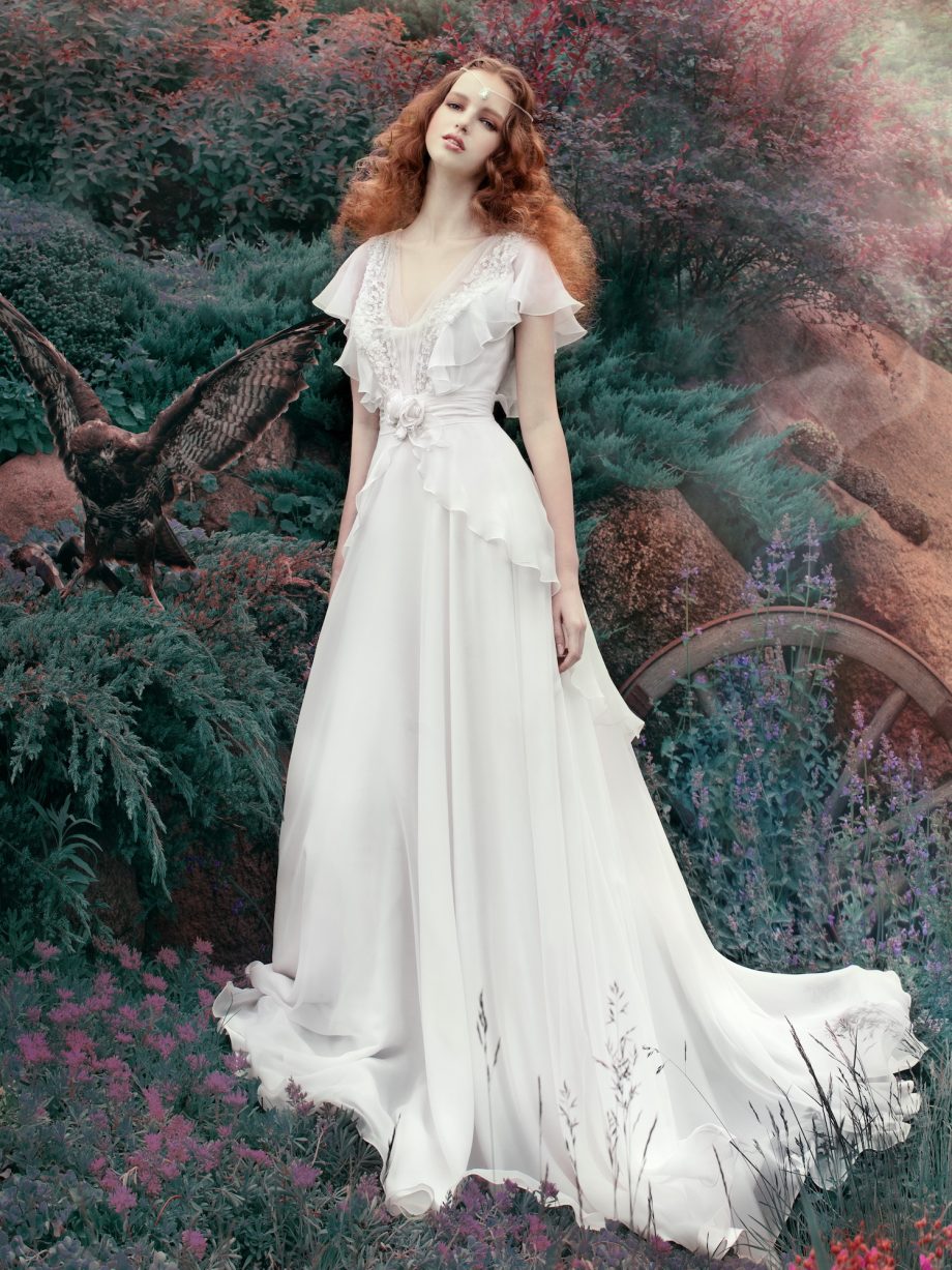 A bride wearing a bohemian sheath wedding dress with ruffled short sleeves and lace detailing, standing in a dreamy outdoor setting. Perfect for bohemian wedding dresses Toronto.